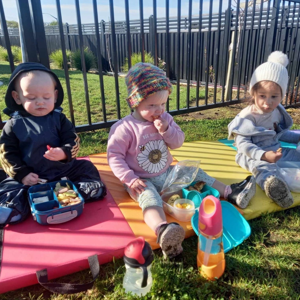 A cozy indoor scene depicting a caregiver gently rocking an infant in a beautifully decorated nursery. The lighting is soft and warm, creating an inviting atmosphere with golden hues, making the space feel safe and nurturing. The room features textured pastel walls, with plush toys scattered around, and a comfortable rocking chair, adorned with a patterned cushion. The caregiver, a diverse individual with a warm smile, wears a colorful apron, reflecting the joy and care given to the baby. In the background, a large window lets in natural light, illuminating the room, while soft curtains flutter gently. The floor is covered with a soft rug, enhancing the comforting vibe. Shot from a slightly high angle, emphasizing the emotional connection between caregiver and infant, with an 8K resolution and hyperrealistic detail, in the style of Anne Geddes.